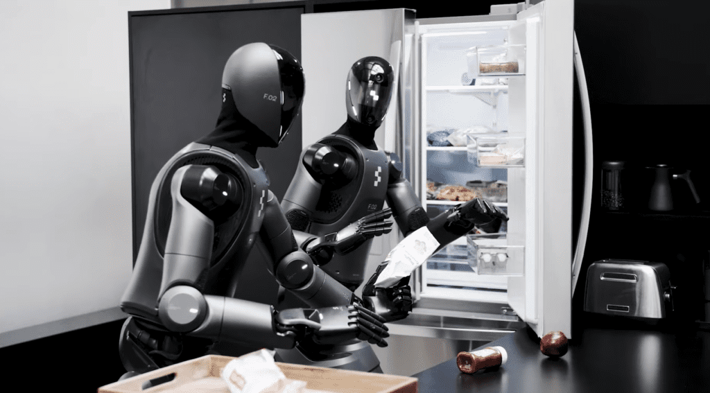 Dos robots humanoides de Figure AI colaboran en una cocina, manipulando alimentos frente a un refrigerador abierto, demostrando interacción autónoma avanzada.