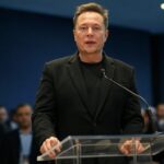 Elon Musk en un evento oficial, vestido de negro, hablando frente a un micrófono y un público atento, durante la presentación de Grok 3.